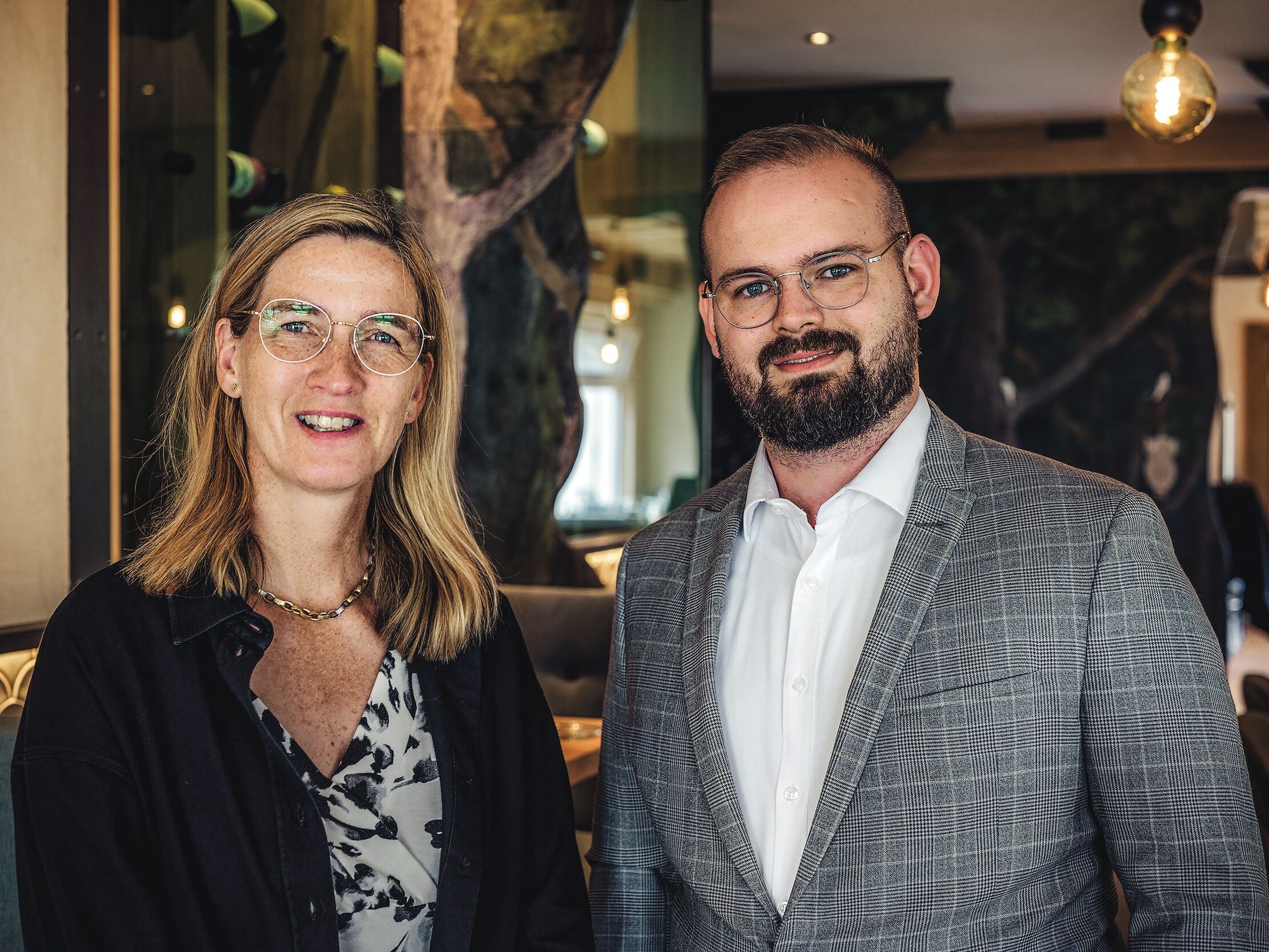 Ein Mann und eine Frau stehen zusammen, beide lächeln, der Mann trägt einen Anzug und eine Brille.